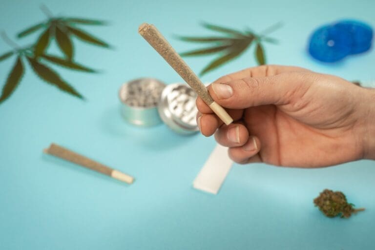 A man holding a preroll with a blurred grinder and cannabis leaves in the background.